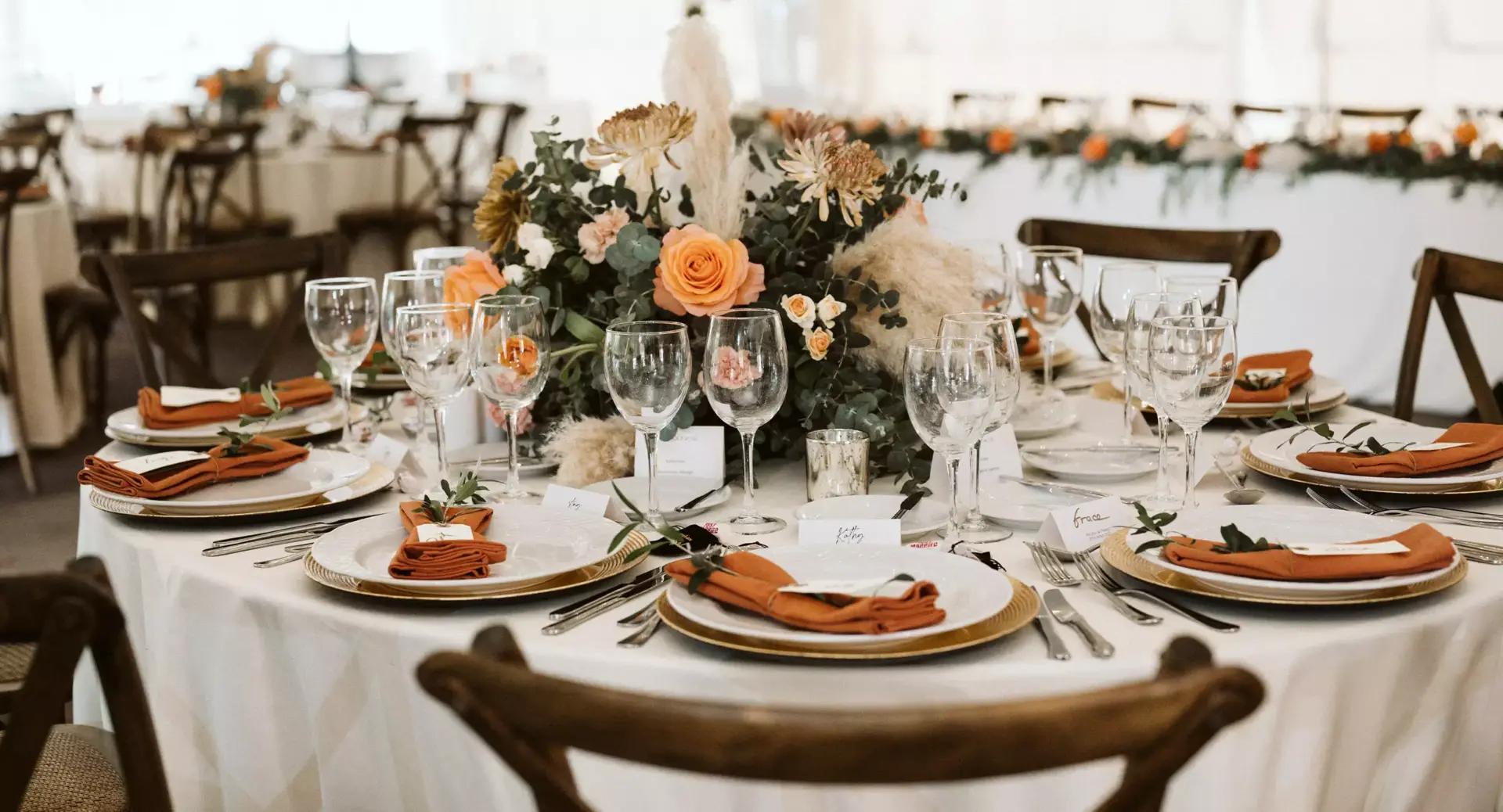 A round banquet table is set with plates, glasses, orange napkins, name cards, and a floral centerpiece with orange and cream flowers.