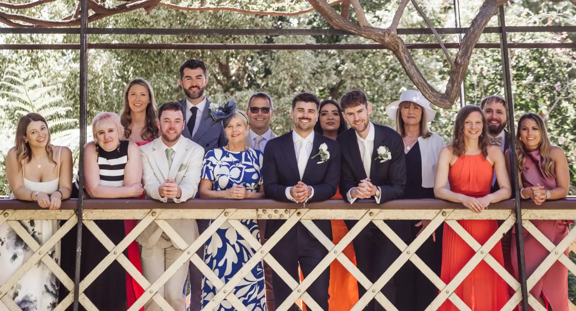 Group of people dressed formally, standing and leaning on a wooden railing outdoors, posing for a photo with greenery in the background.