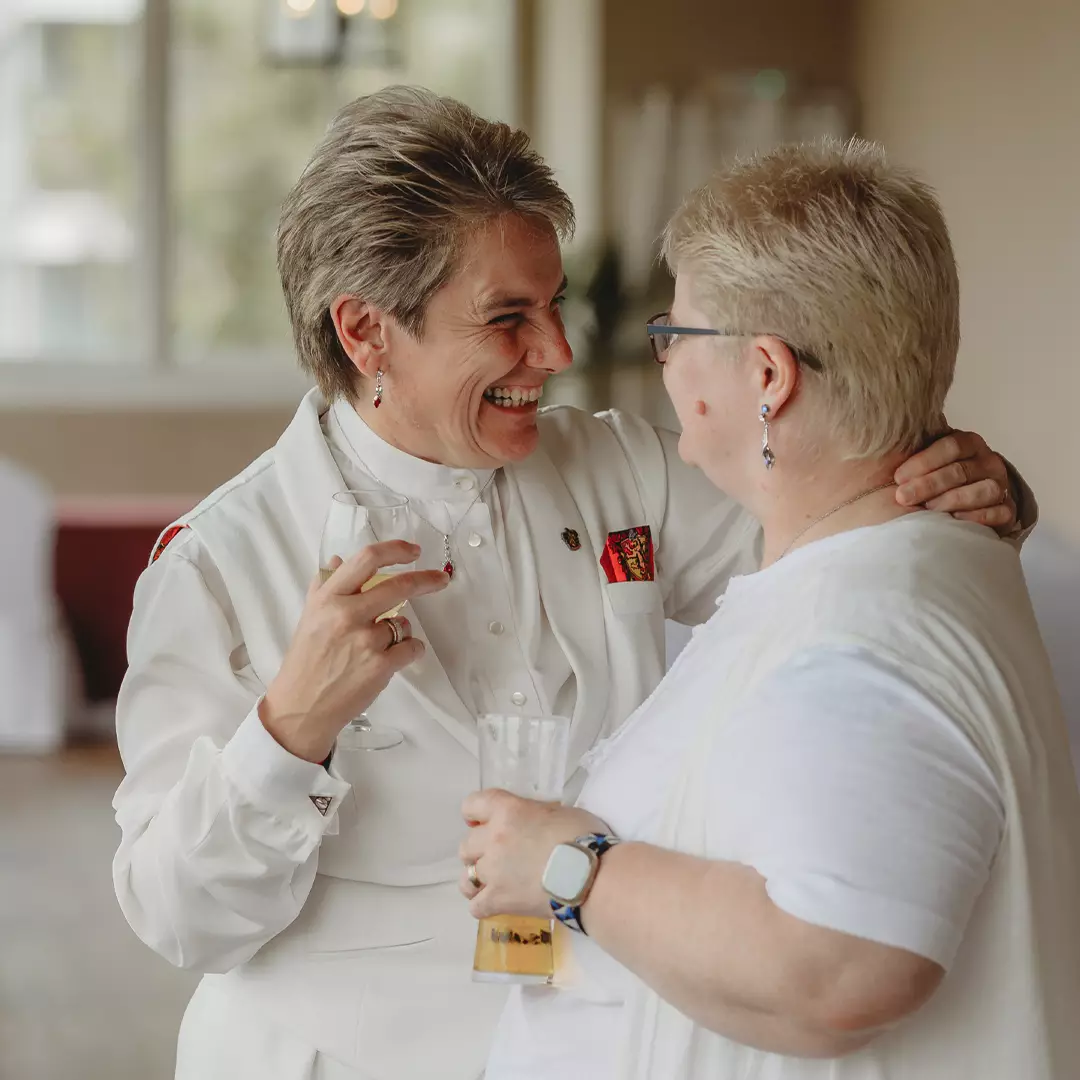 Two women in white outfits smile and chat while holding drinks at an indoor event.