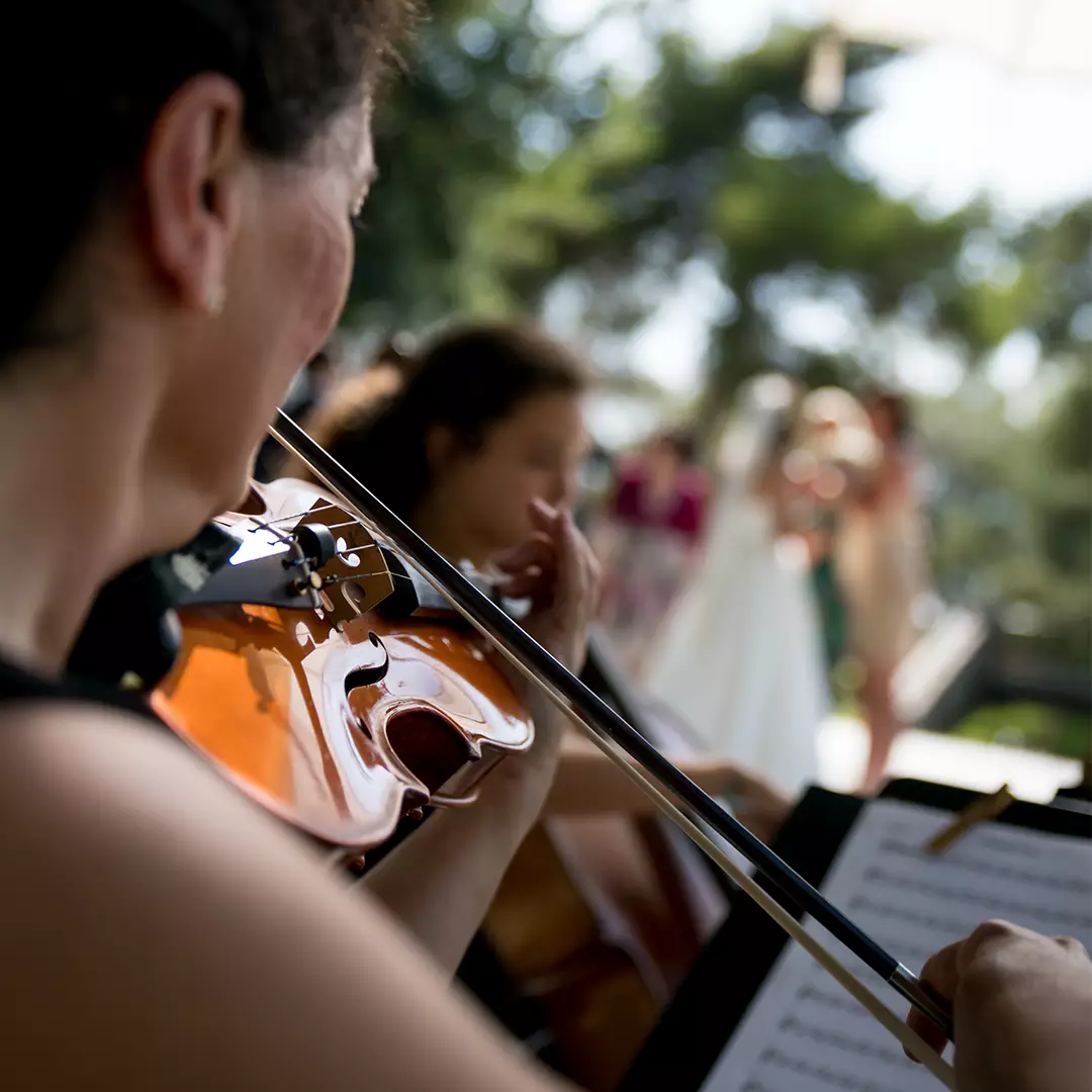 A violinist plays music outdoors with sheet music in front, while other musicians and blurred figures are visible in the background.