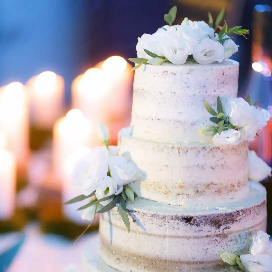 Three-tiered semi-naked cake decorated with white flowers and greenery, with blurred candles glowing in the background.