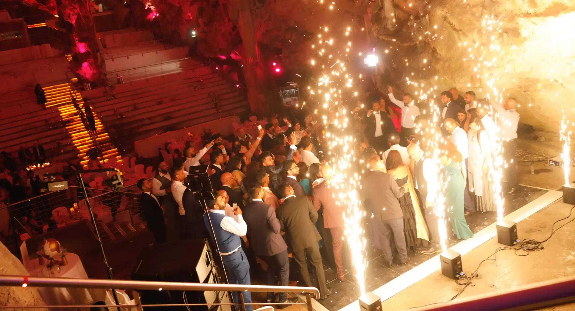 A group of people gathers on a stage with indoor fireworks, surrounded by seated guests in an amphitheater-style venue lit with warm lighting.
