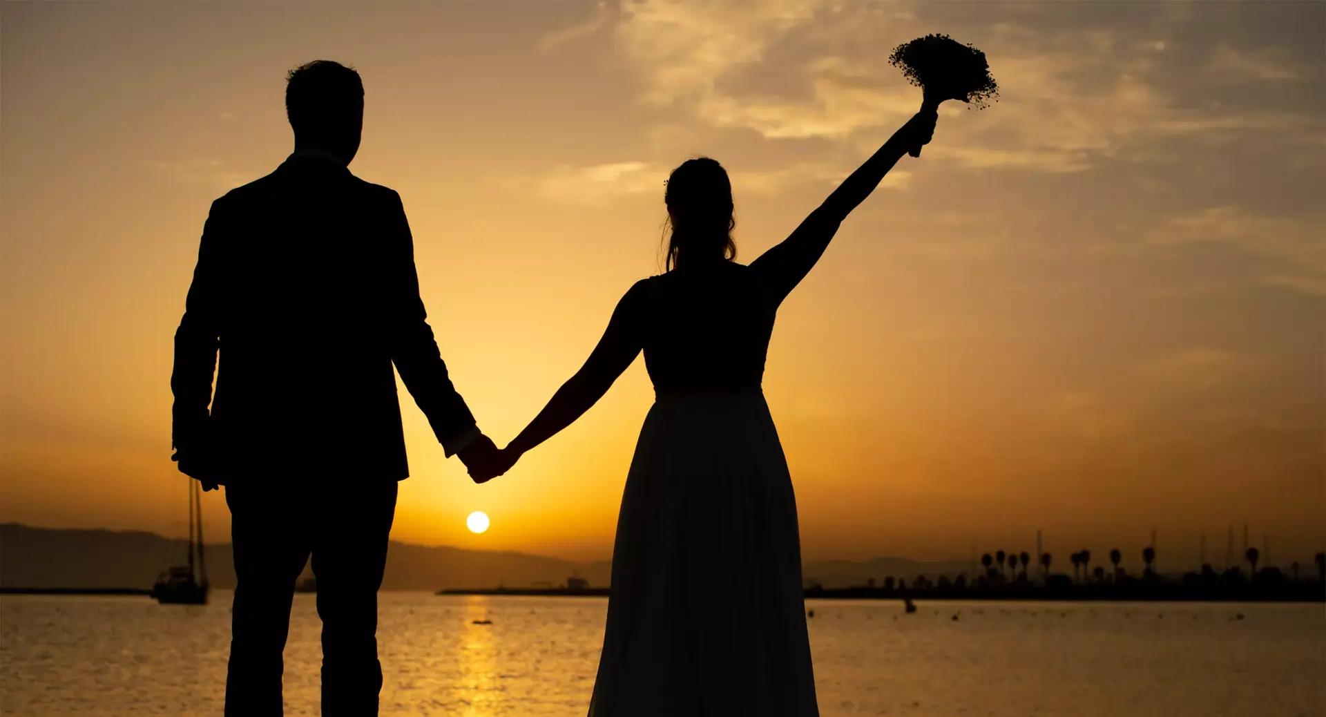 Silhouette of a couple holding hands by the water at sunset, with one person raising a bouquet in the air.
