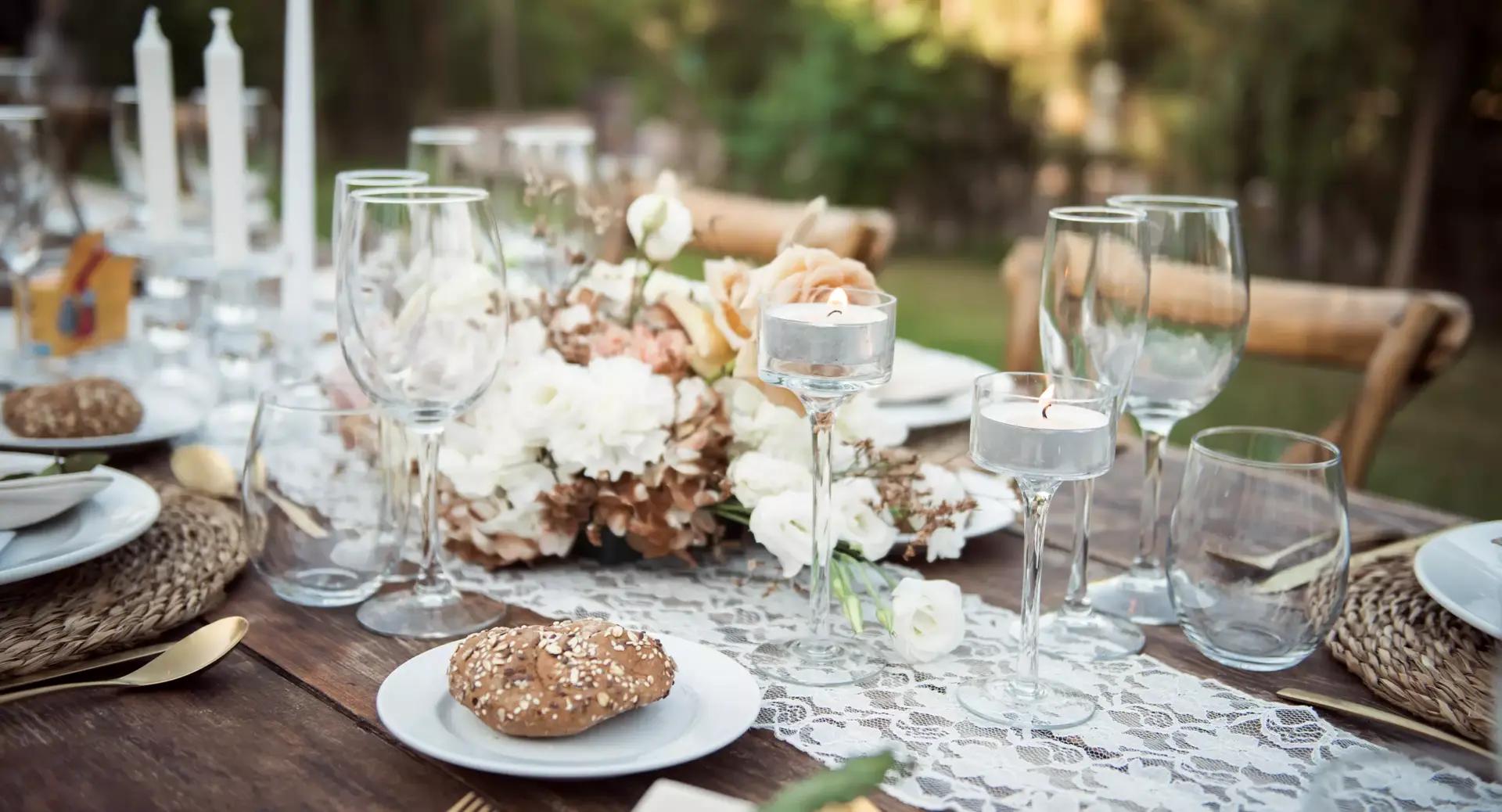 Elegant outdoor table setting with glassware, white candles, rolls on plates, and a floral centerpiece, arranged on a wooden table with a lace table runner.