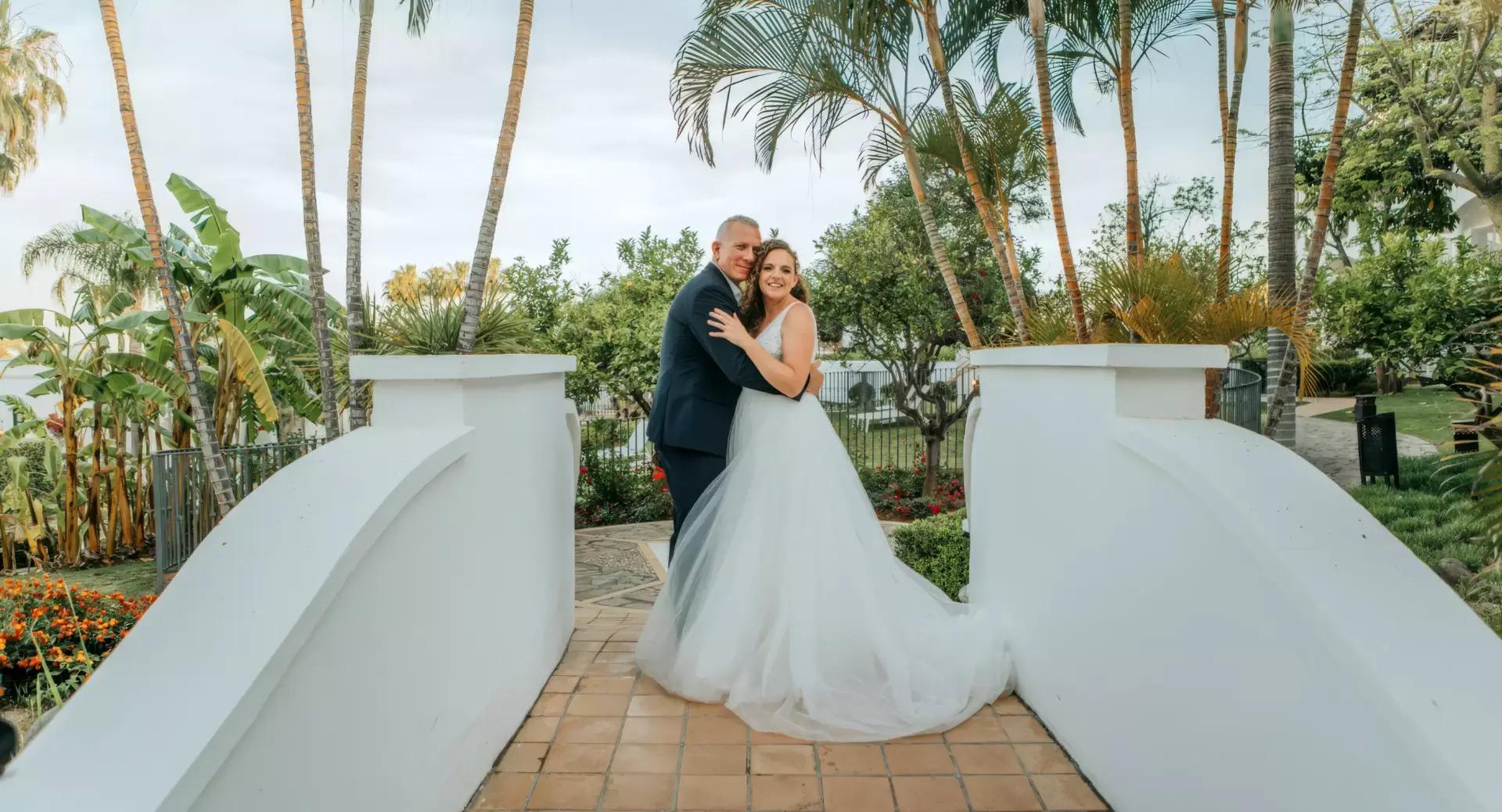 A bride and groom stand embracing on a tiled walkway, surrounded by white walls and tropical plants, posing for a wedding photo outdoors.