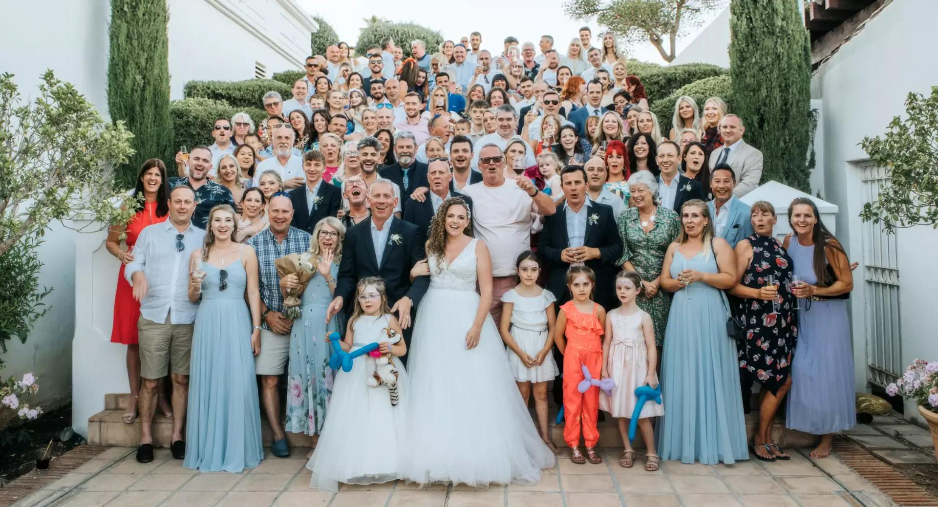 Large group of people, including a bride and groom, pose for a wedding photo outdoors on steps, with bridesmaids in blue dresses and children holding balloon animals.