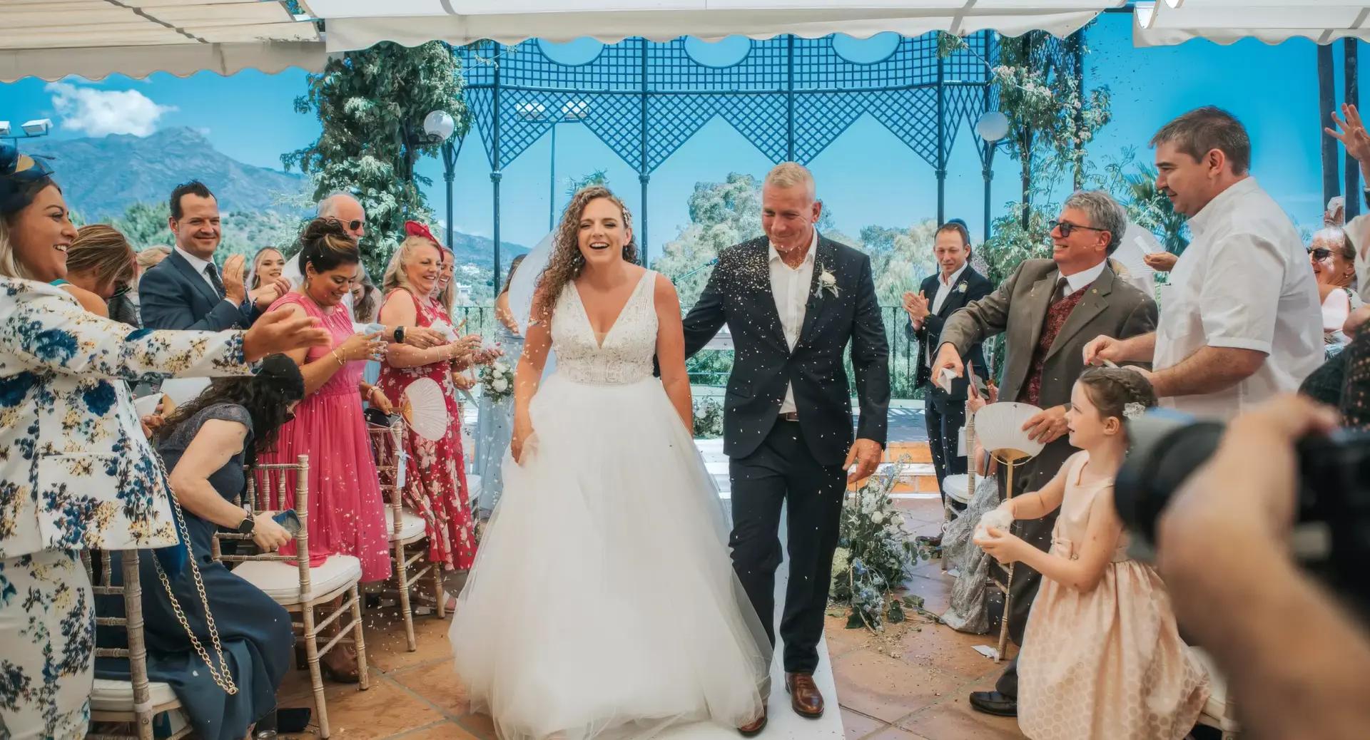 A bride and groom walk down the aisle smiling as guests throw confetti during an outdoor wedding ceremony under a decorated canopy.
